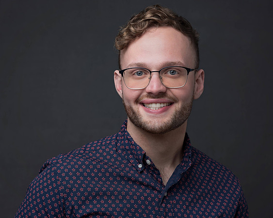 male with curly short hair wearing glasses and a patterned buttondown shirt in a casual headshot