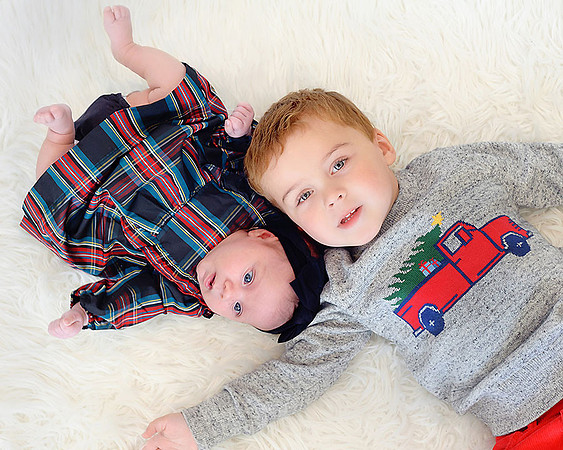 toddler boy wearing Christmas sweater laying on  white fluffy rug with  newborn  sister wearing a plaid dress